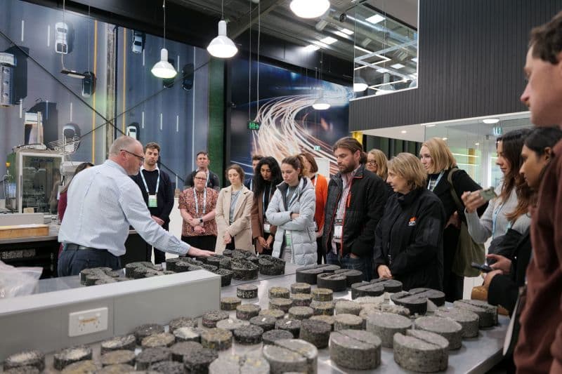 A group of professionals observing a demonstration of road surface materials in a laboratory setting.